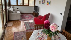 A living room with a sofa, red chair, and a table with a floral tablecloth. There are rugs on the floor and a TV on a stand. - Casa Malù----------- (Messina)