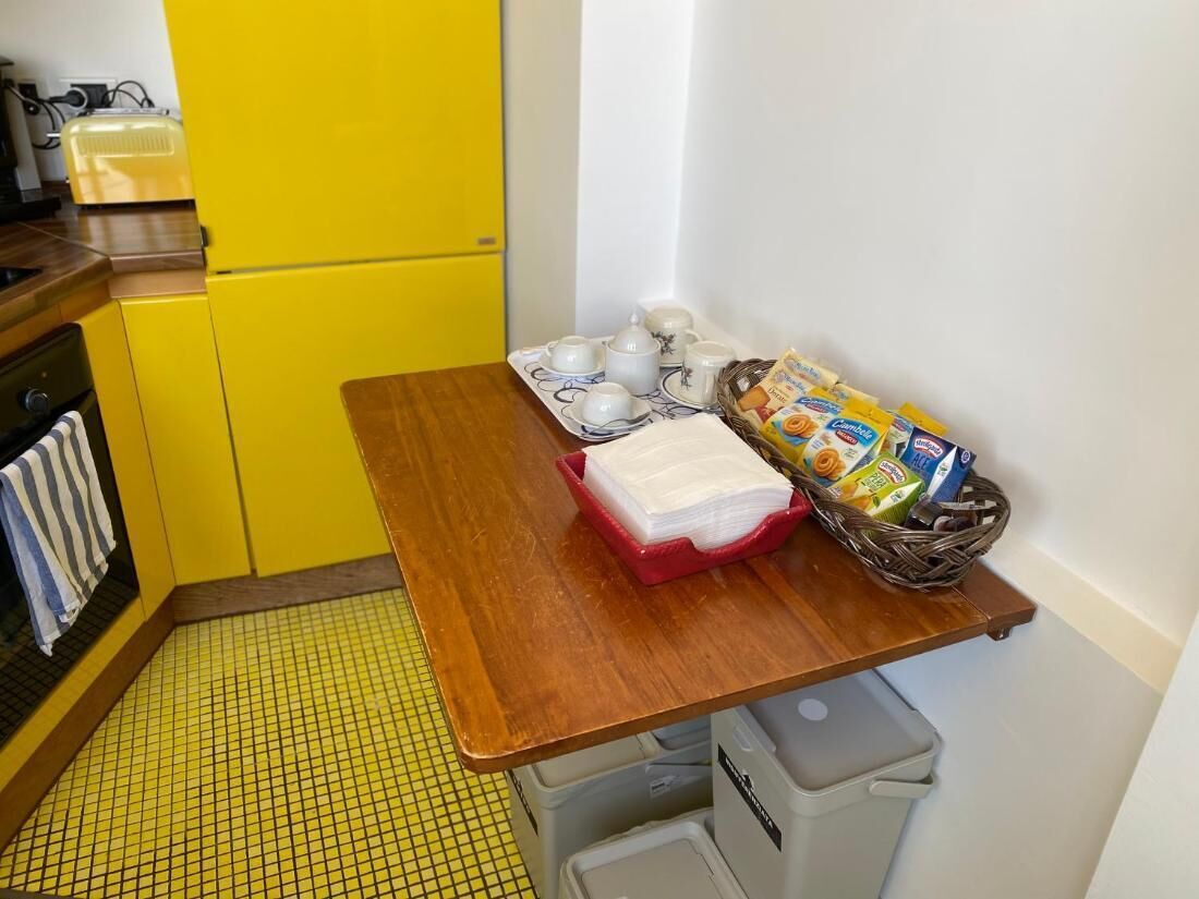 A small kitchen with a yellow refrigerator, a wooden table, and a sink. There are white cups and a basket of bread on the table.
