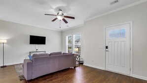 Un salon avec un canapé gris, un ventilateur de plafond, un téléviseur à écran plat et une porte dotée de panneaux en verre dépoli.