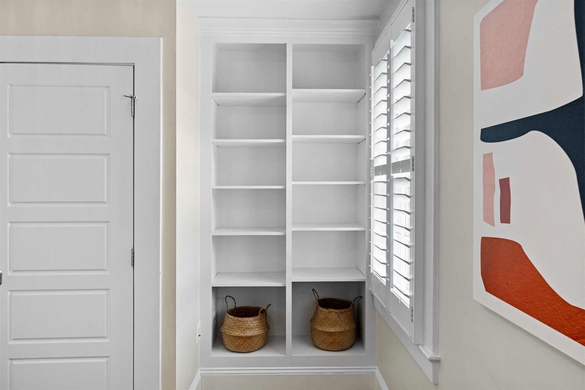 A white interior space with a door, a built-in shelving unit, and a window with blinds.