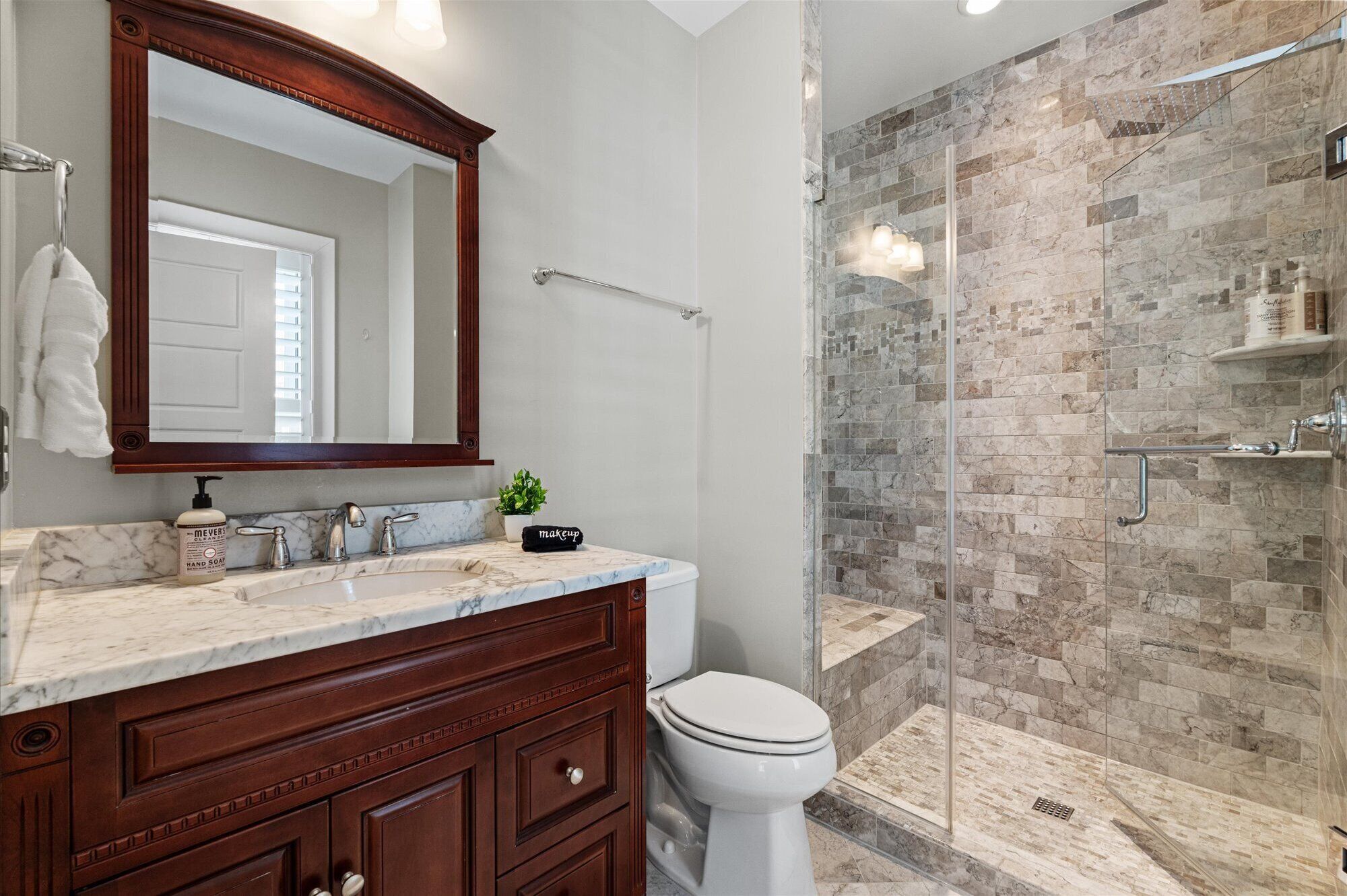 A bathroom with a marble vanity, a large mirror, a toilet, and a glass-enclosed shower with stone tiles.