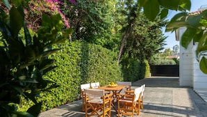 Een terras buiten met een houten tafel en stoelen, omringd door weelderige begroeiing en een haag.