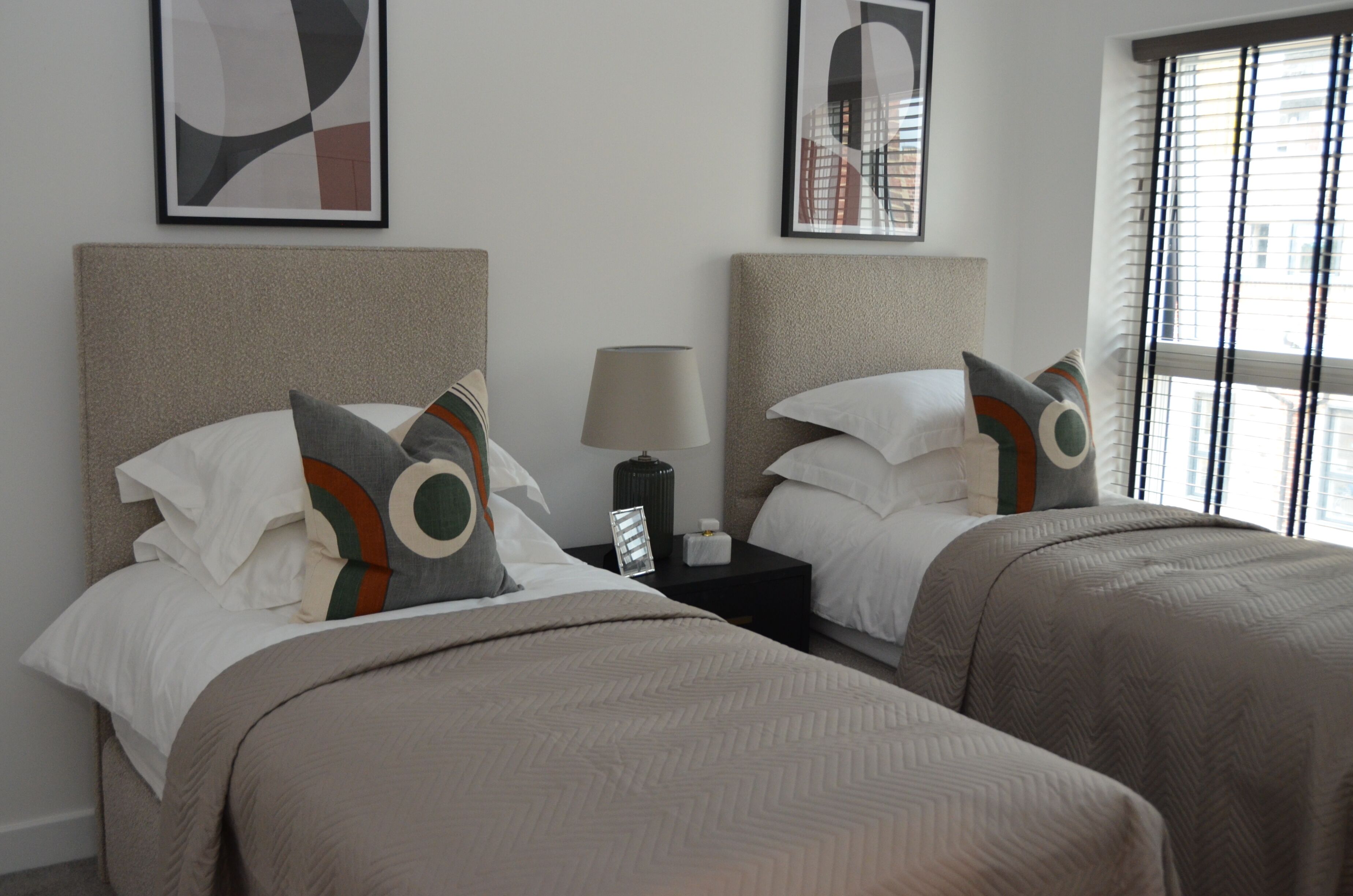 A hotel room with two beds, each with a headboard, a bedside table with a lamp, and a window with blinds.