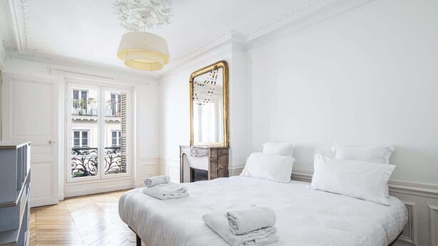 A hotel room with a large bed, white bedding, a mirror, and a chandelier.