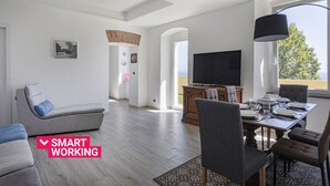 Una sala moderna con un sofá gris, una mesa de comedor con sillas, un televisor de pantalla plana y un amplio ventanal con vistas a zonas verdes.