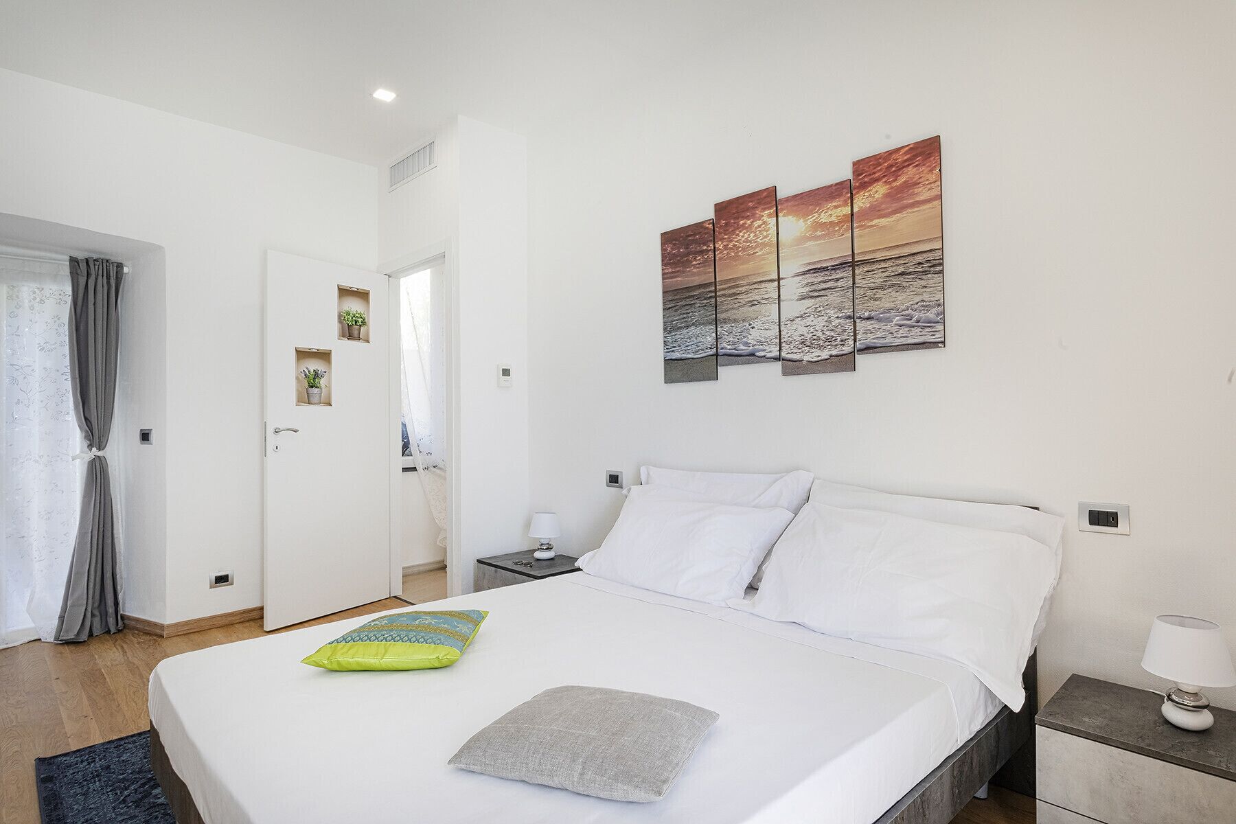 A hotel room with a bed, a nightstand, a lamp, and a painting of a sunset over the sea.