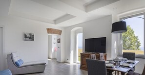 Un salon moderne avec un canapé gris, une table à manger avec des chaises, un téléviseur à écran plat et une grande fenêtre donnant sur un paysage verdoyant.