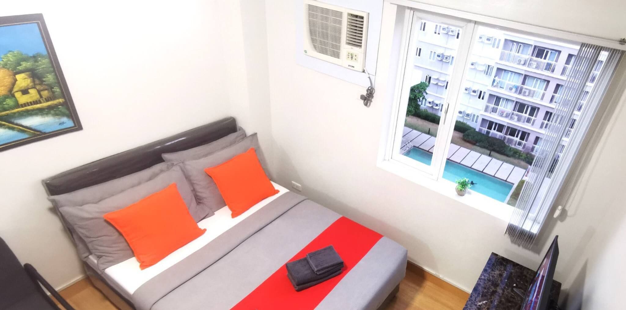 A neatly made bed with grey and orange bedding, a small bedside table with a remote control, and a window with a view of a building and a pool.