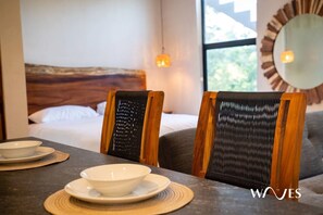 A hotel room with a bed, two chairs, a dining table with plates and bowls, a wall-mounted mirror, and a window with natural light.