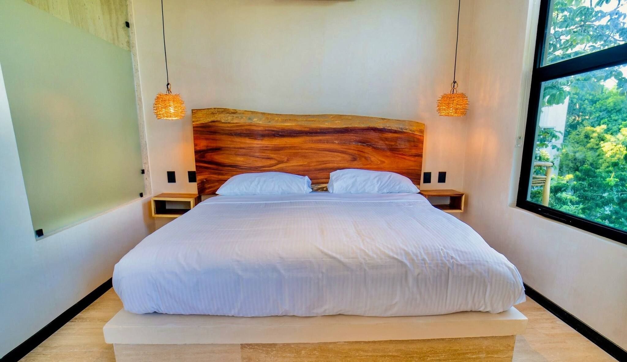 A modern bedroom with a large bed, wooden headboard, two bedside tables, and a view of greenery through a large window.