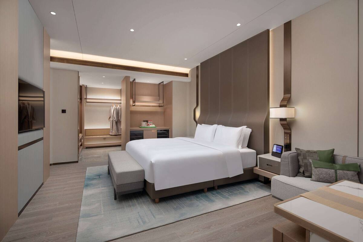 A modern hotel room with a large bed, bedside tables, a sofa, and a desk with a lamp.