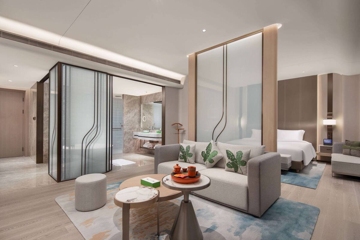 A modern hotel room with a sofa, a bed, a small table, and a bathroom visible through a glass partition.