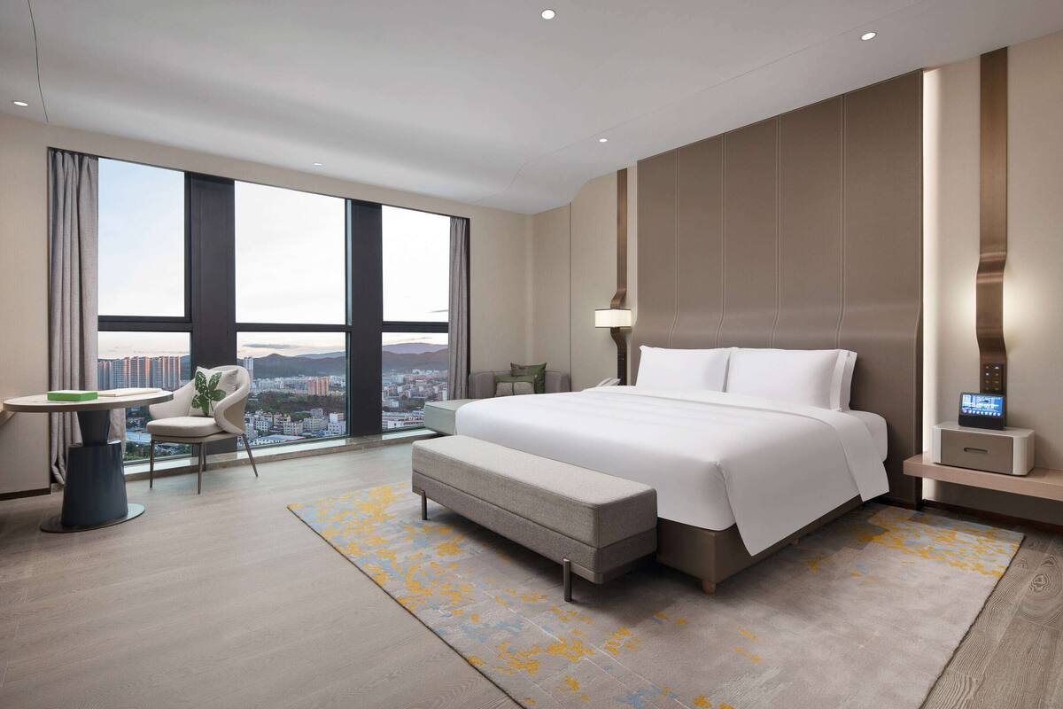 A modern hotel room with a large bed, a desk, a chair, and a view of the city.