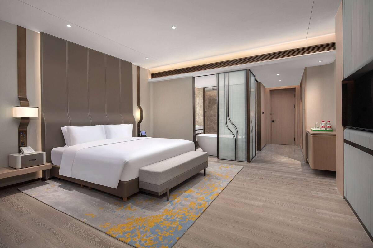 A modern hotel room with a large bed, bedside table, lamp, and a bathroom with a bathtub and shower.