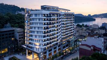 Un hotel de varios pisos con diseño moderno, con balcones en cada planta, situado cerca de una masa de agua y rodeado de vegetación.