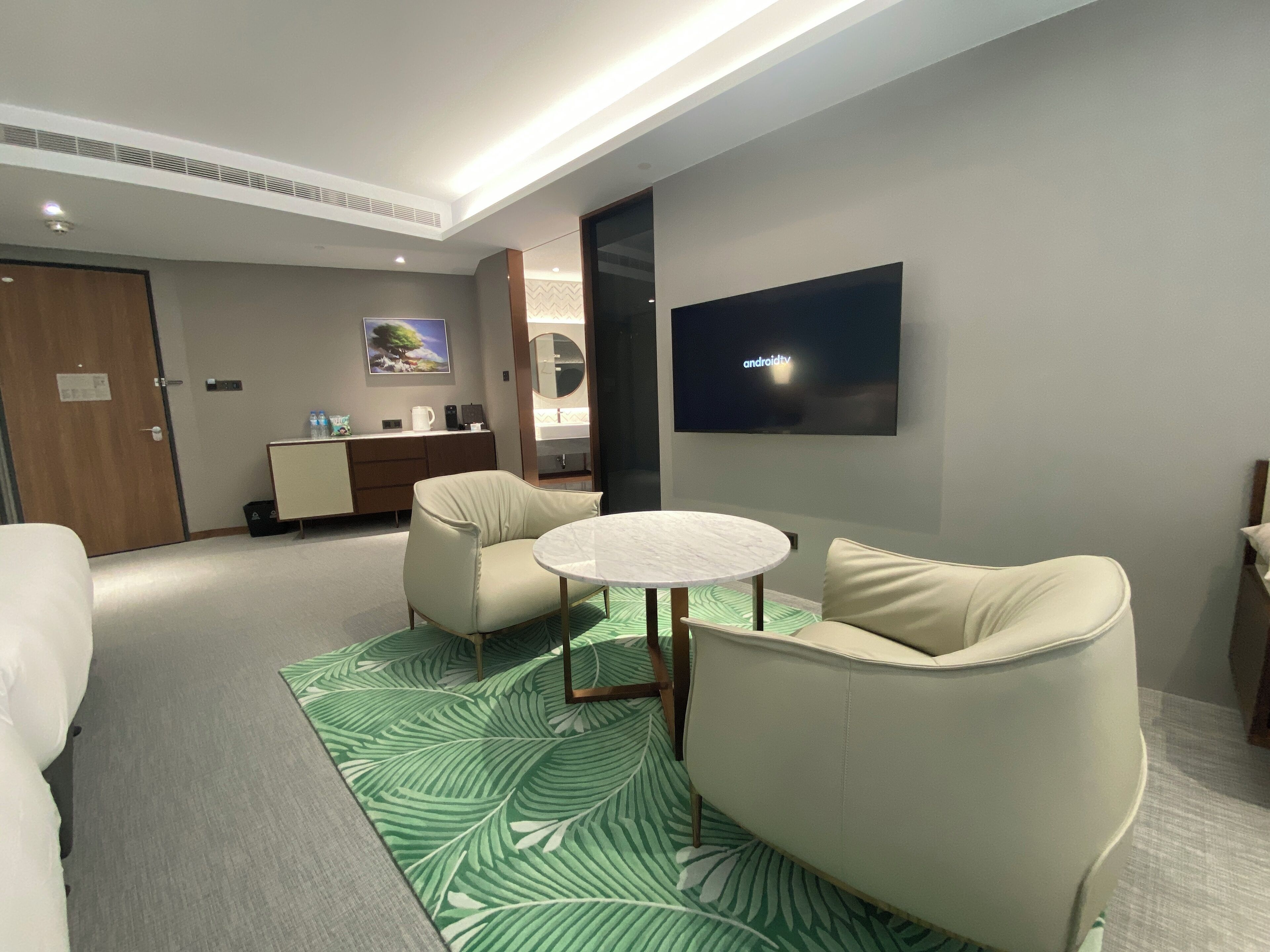A modern hotel room with a round table, two armchairs, a flat-screen TV, a minibar, and a large window.
