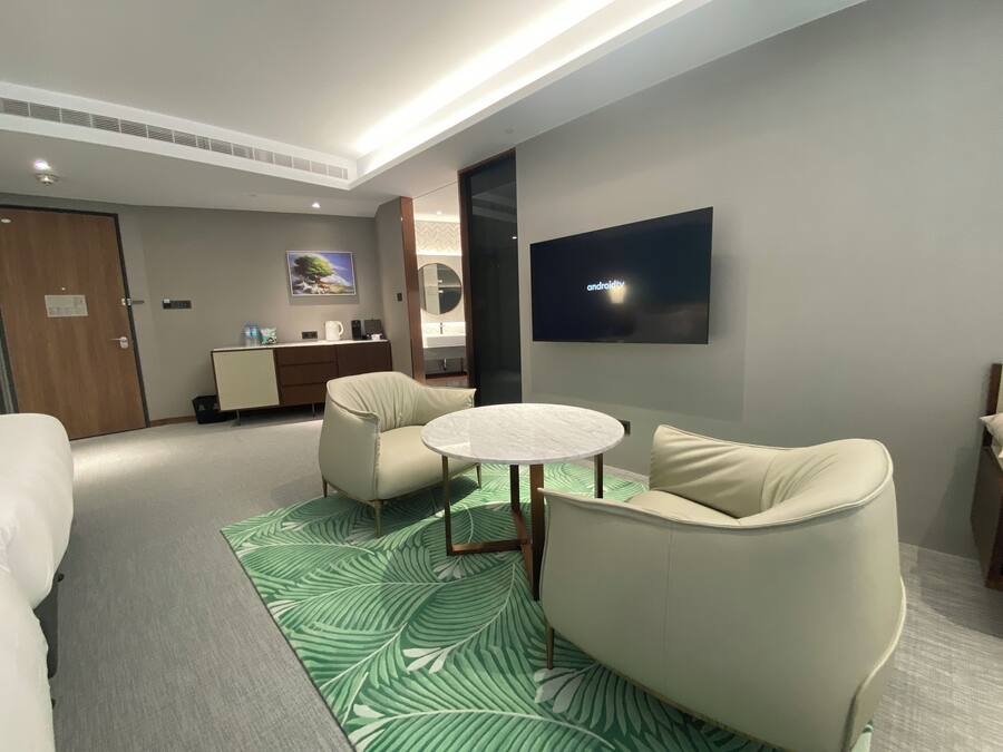 A modern hotel room with a round table, two armchairs, a flat-screen TV, a minibar, and a large window.