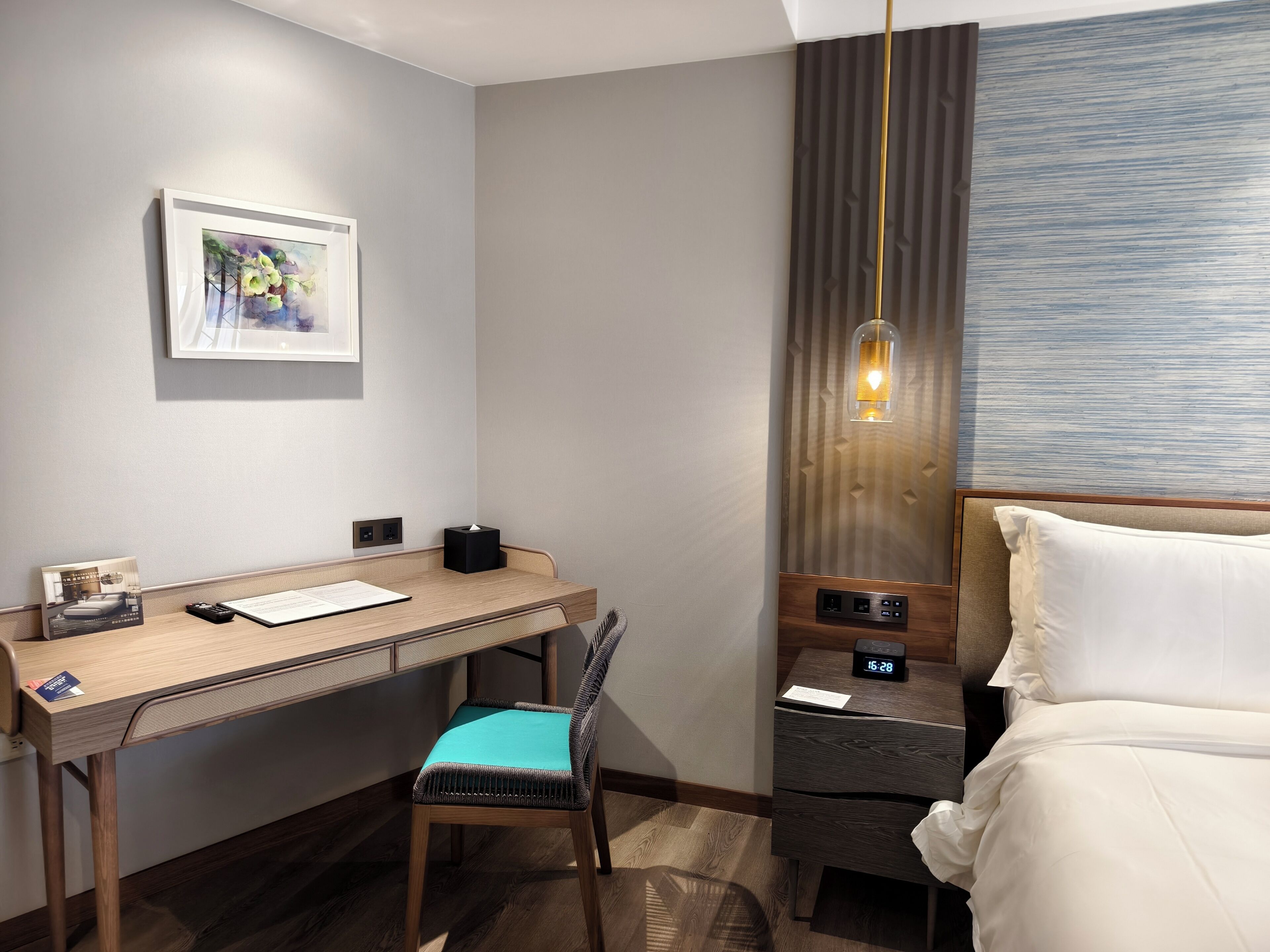 A hotel room with a desk, chair, bed, bedside table, and a wall-mounted lamp.