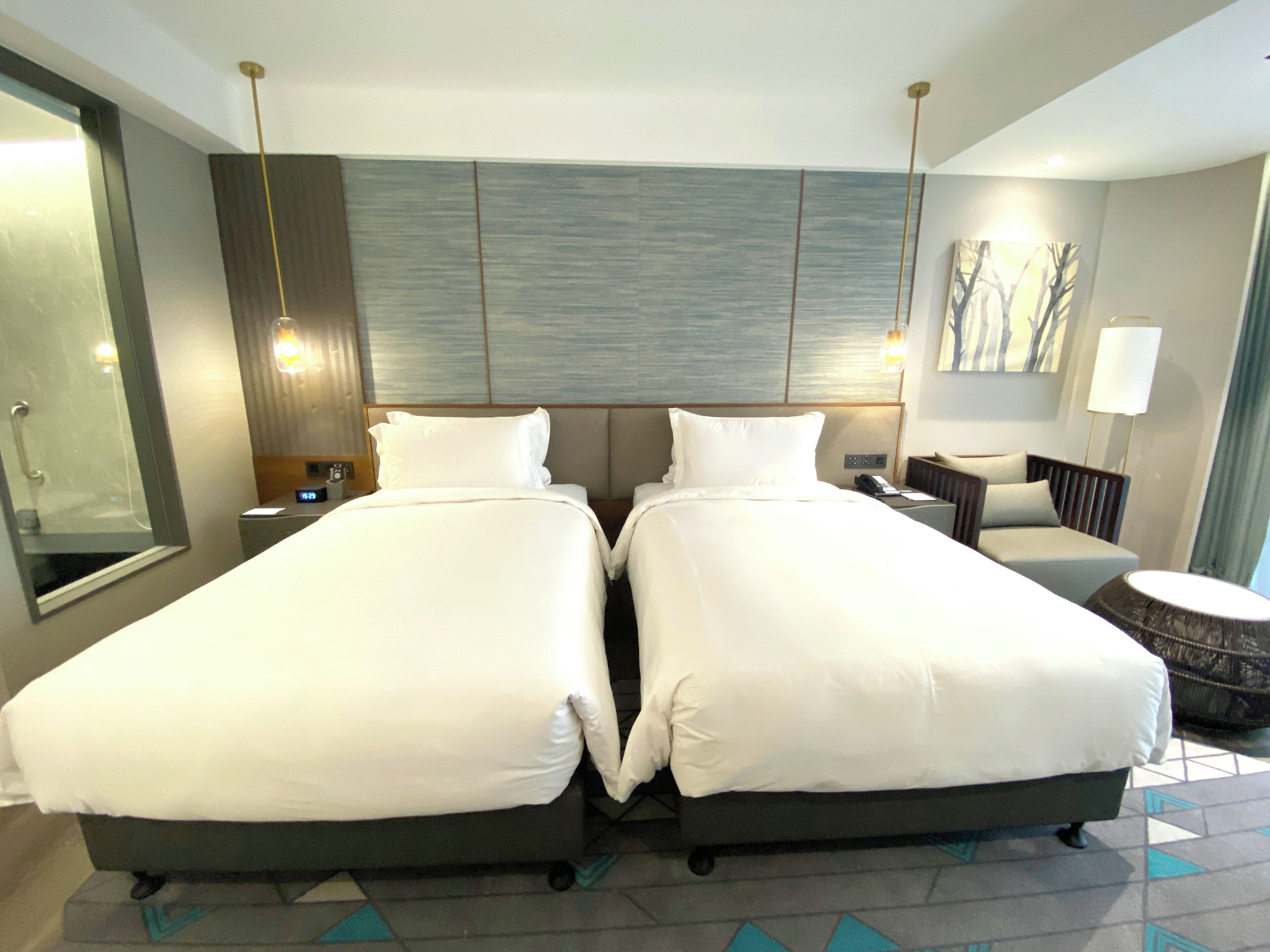 A hotel room with two beds, a desk, a chair, a lamp, and a painting on the wall.