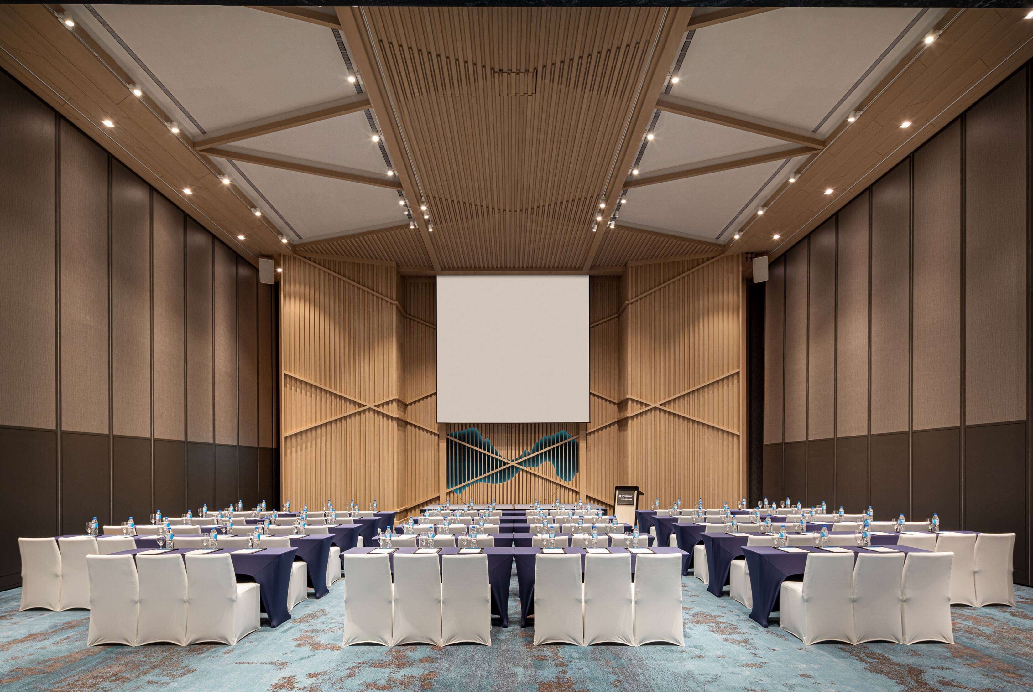 A large conference room with rows of tables and chairs, a large screen at the front, and a wooden ceiling design.