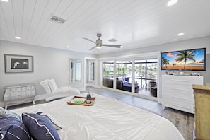 Une chambre avec un grand lit, un ventilateur de plafond, une télévision et une vue sur la plage par la fenêtre.
