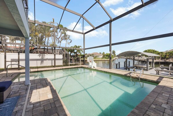 A pool area with a clear view of the water, a covered seating area, and a dock extending into a body of water.