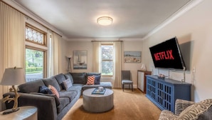 Ein Wohnzimmer mit einem grauen Sofa, einem runden Couchtisch, einem blauen Schrank und einem Fernseher, der 'NETFLIX' anzeigt.
