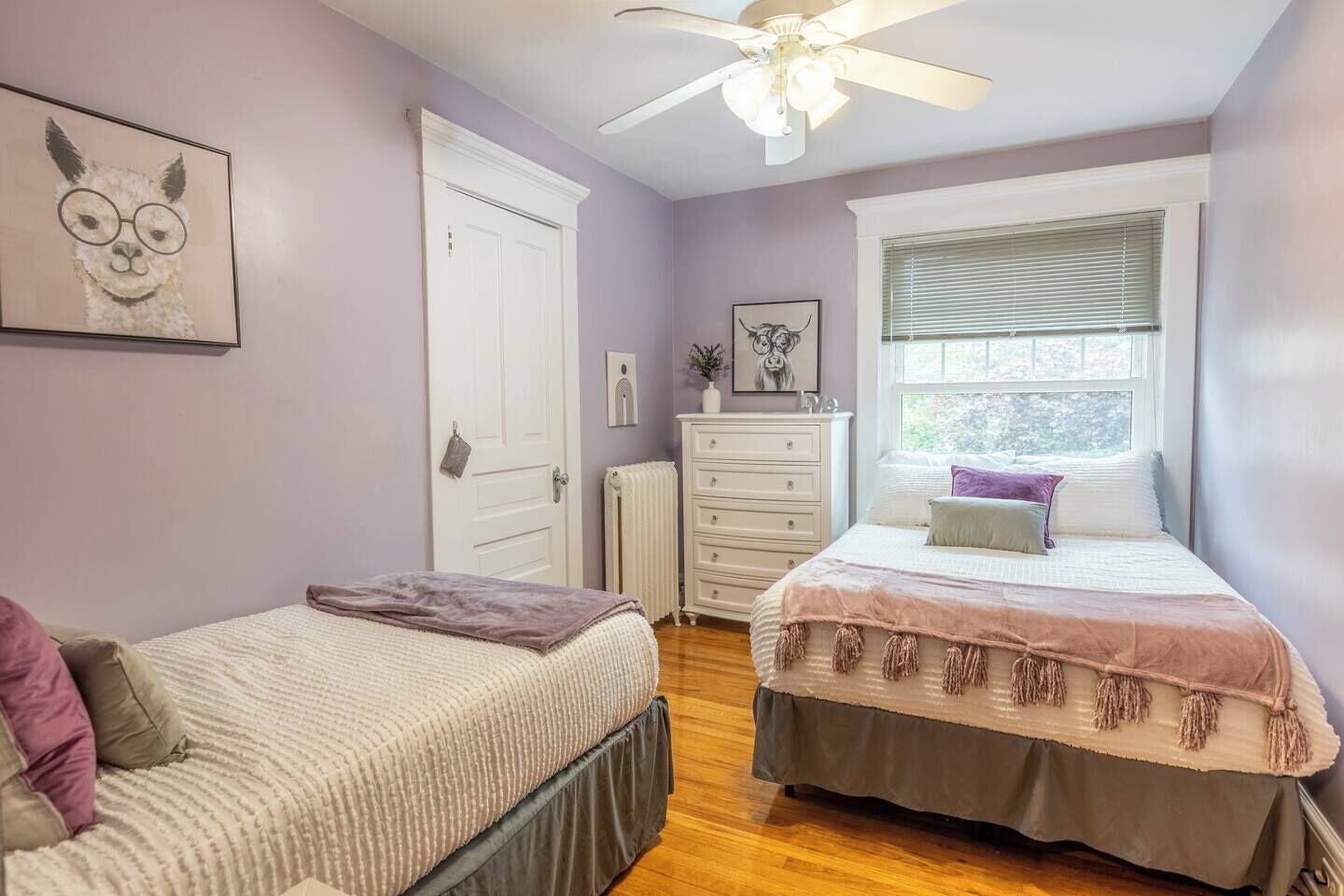 Une chambre avec deux lits, un ventilateur de plafond, une fenêtre avec des stores et un radiateur.