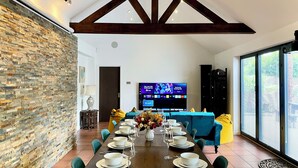 A dining area with a long table set for six, a stone wall, a blue sofa, and a TV displaying a gaming channel.