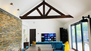 A spacious living room with a high ceiling, exposed wooden beams, a stone wall, a large flat-screen TV, and a sliding glass door leading to an outdoor area.
