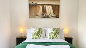 A neatly made bed with a green blanket, white pillows, and a green leaf-patterned pillow. There are two bedside lamps on either side of the bed.