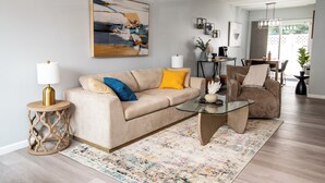 Un salon avec un canapé beige, une table basse en verre et un tapis à motifs.