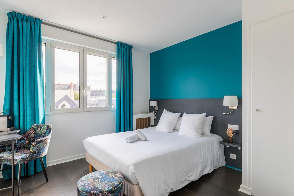 A modern hotel room with a large bed, a desk with a chair, and a view of the city through a window.