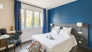 A hotel room with a large bed, a desk with a chair, a window with curtains, and a view of buildings outside. - Hôtel Restaurant Le Victoria (Rennes)
