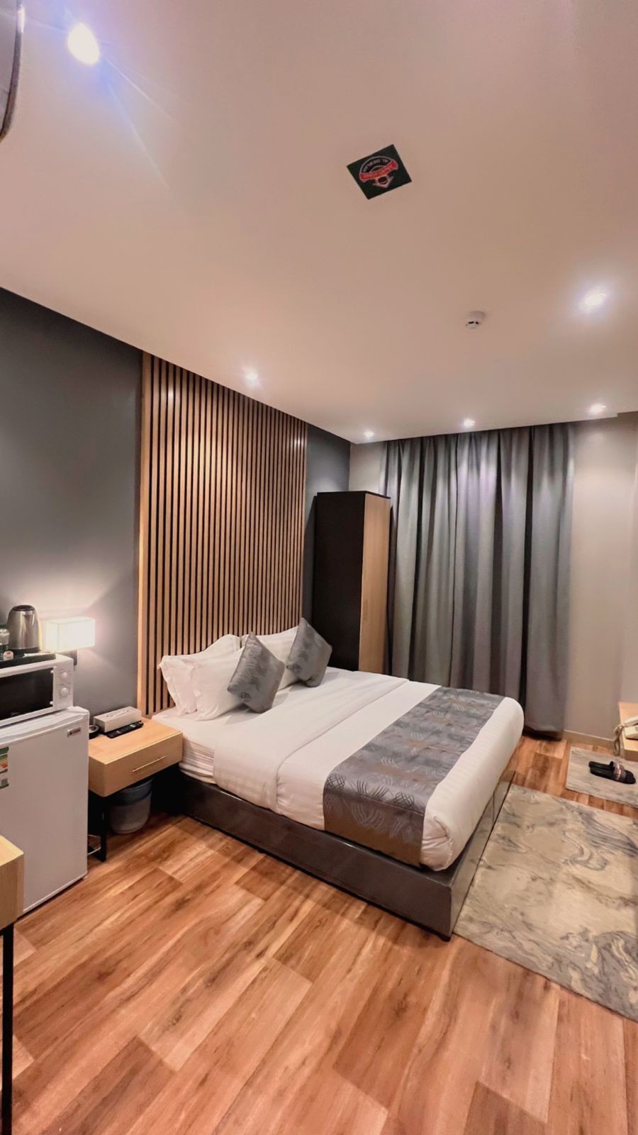 Quarto de hotel moderno com piso de madeira, cama com colcha cinza e branca, mesa de cabeceira com luminária, pequena mesa com chaleira e guarda-roupa de cor escura.