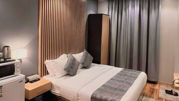 Quarto de hotel moderno com piso de madeira, cama com colcha cinza e branca, mesa de cabeceira com luminária, pequena mesa com chaleira e guarda-roupa de cor escura.