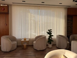 A modern living room with a large window covered by vertical blinds, two armchairs, a small side table with a lamp, and a potted plant. - السعاده سويت للشقق المخدومه (Riyadh)