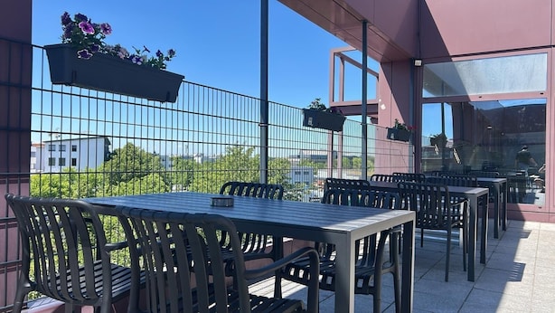 An outdoor patio with tables and chairs, a metal fence, and hanging flower baskets. - Sitya Hostel München (Munich)