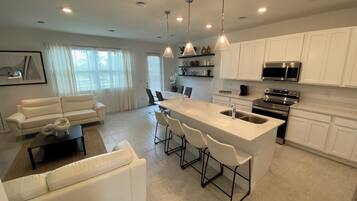 A modern kitchen with white cabinetry, a central island with bar stools, a dining area with a white sofa, and pendant lighting.