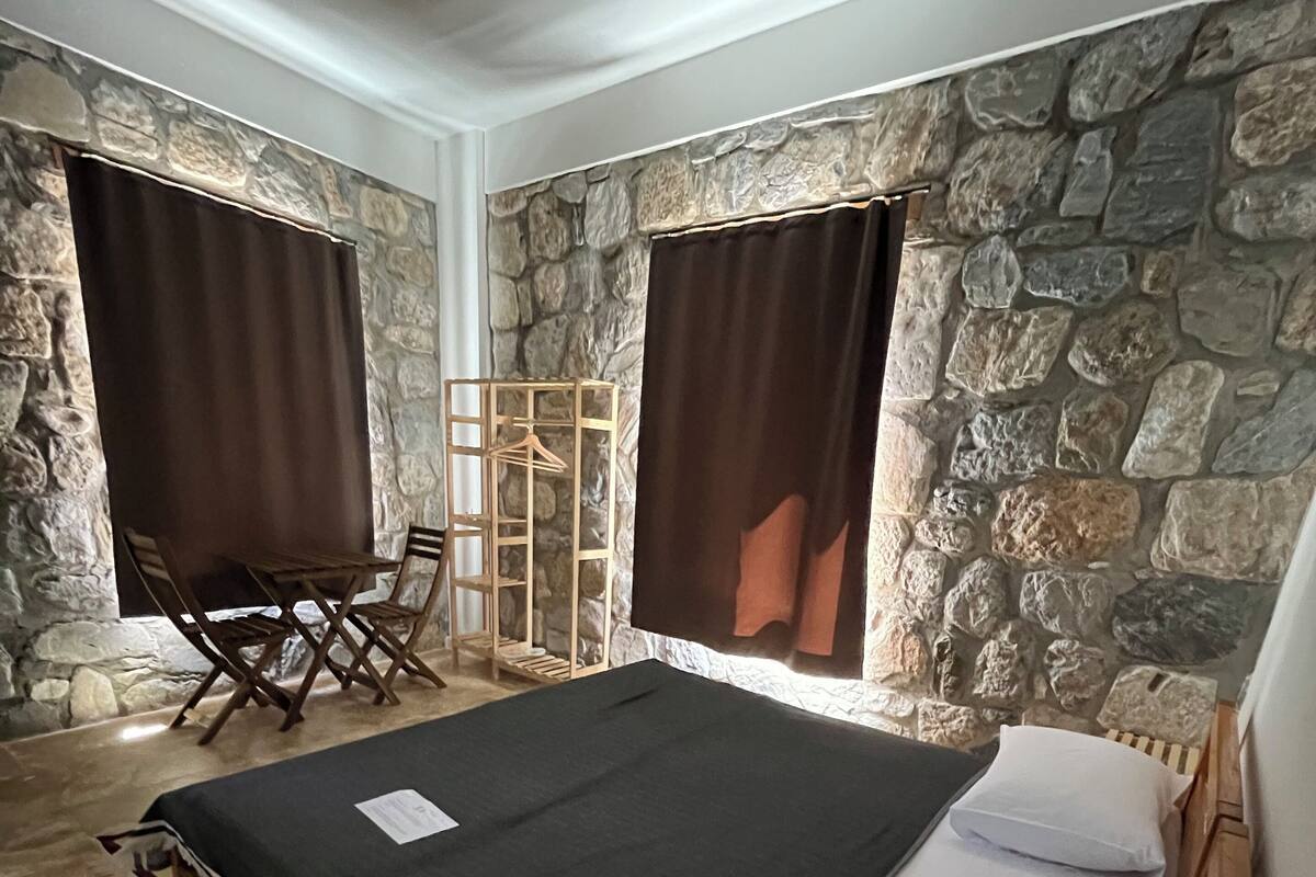 A room with a bed, a wooden chair, a small table, and a clothing rack against a stone wall.