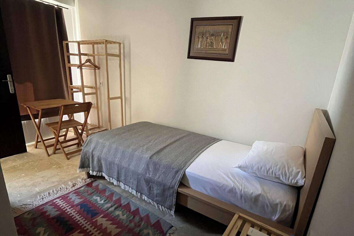A small, neatly arranged bedroom with a bed, a wooden side table, a chair, a hanging rack, and a patterned rug.