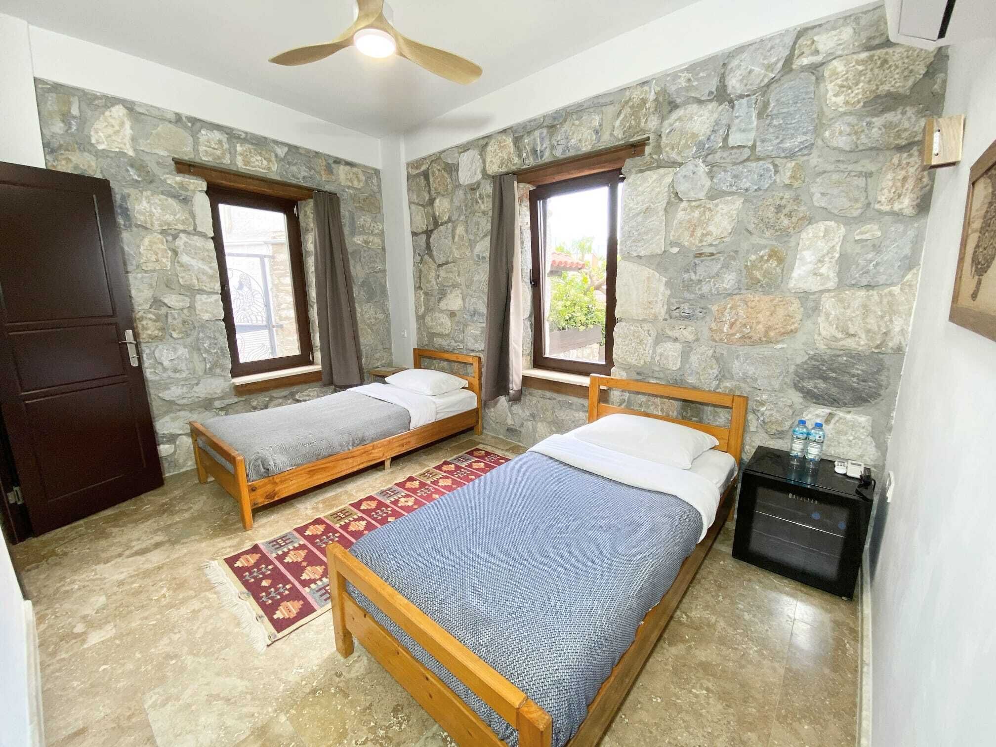 A room with two wooden beds, a ceiling fan, stone walls, and a small table with water bottles.