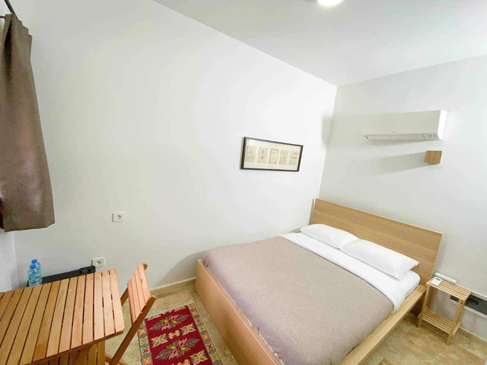 Ein Schlafzimmer mit einem Bett, einem Holztisch, einem Stuhl, einem roten Teppich mit Muster und einer Wand-Klimaanlage.
