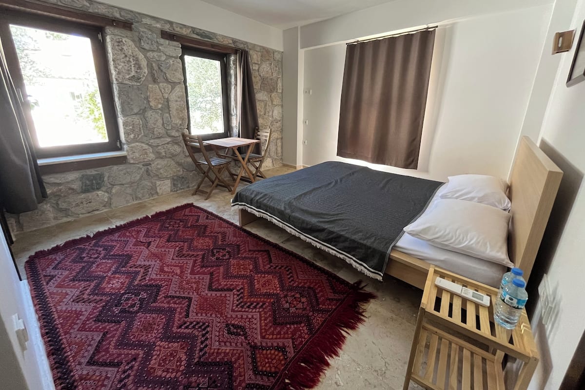 A bedroom with a bed, a wooden nightstand, a small table with chairs, a stone wall, and a large window.