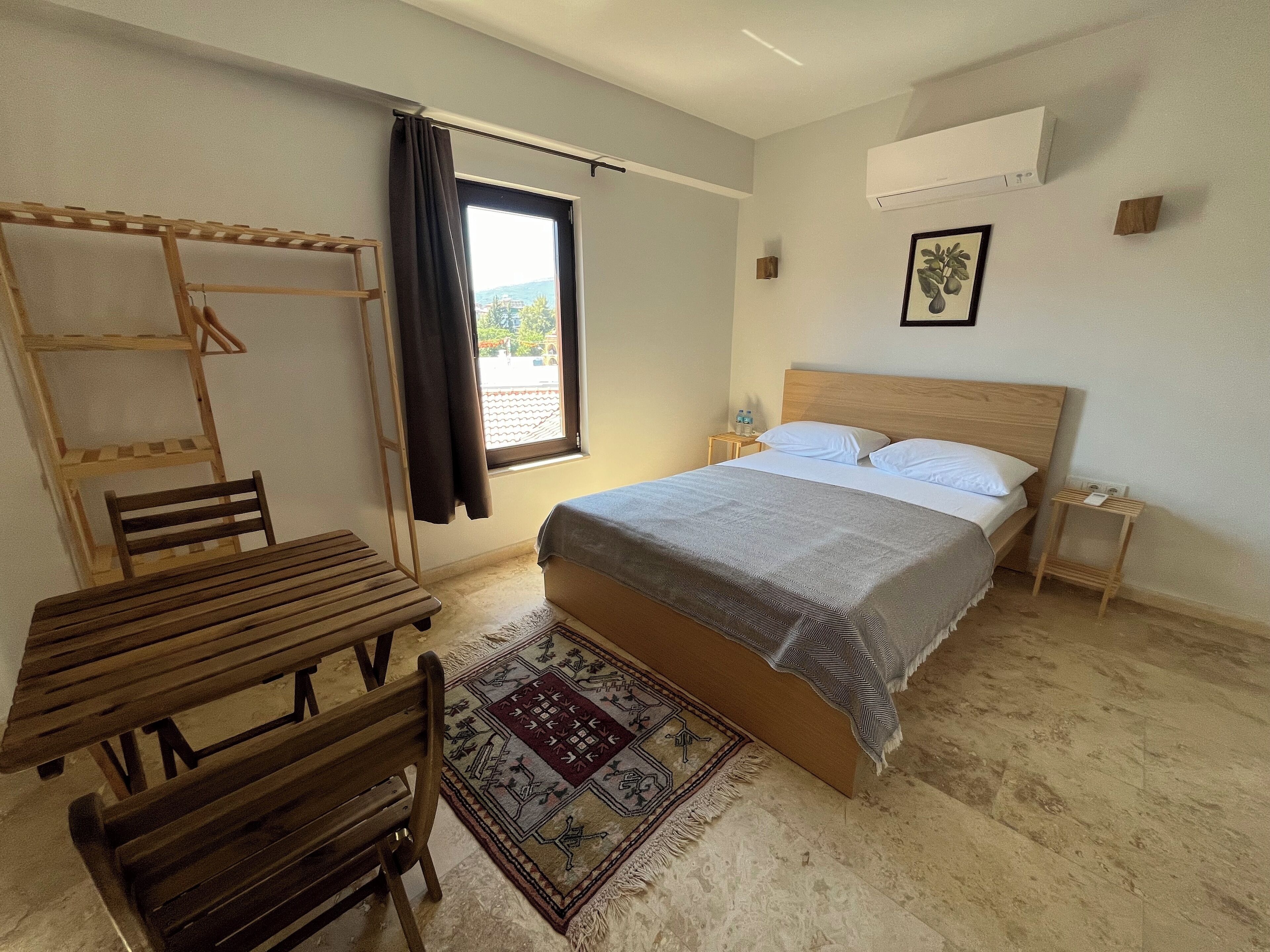Un dormitorio con cama, una mesa de madera con sillas, un ventanal con cortinas, una mesita de noche con lámpara y un aire acondicionado instalado en la pared.