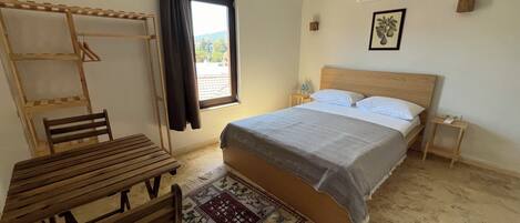 Un dormitorio con cama, una mesa de madera con sillas, un ventanal con cortinas, una mesita de noche con lámpara y un aire acondicionado instalado en la pared.
