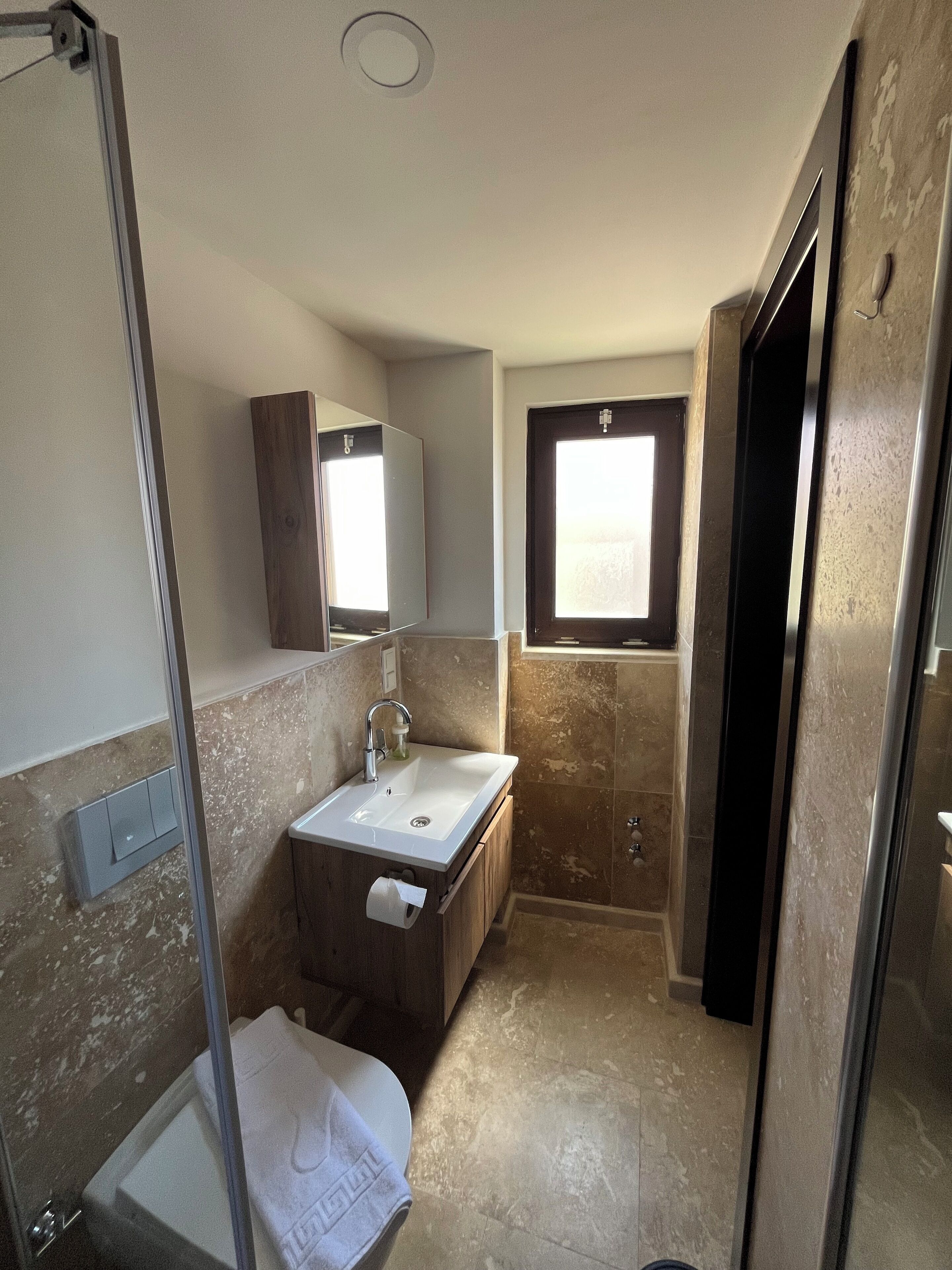 A compact bathroom with a glass shower enclosure, a sink with a mirror, and a window providing natural light.