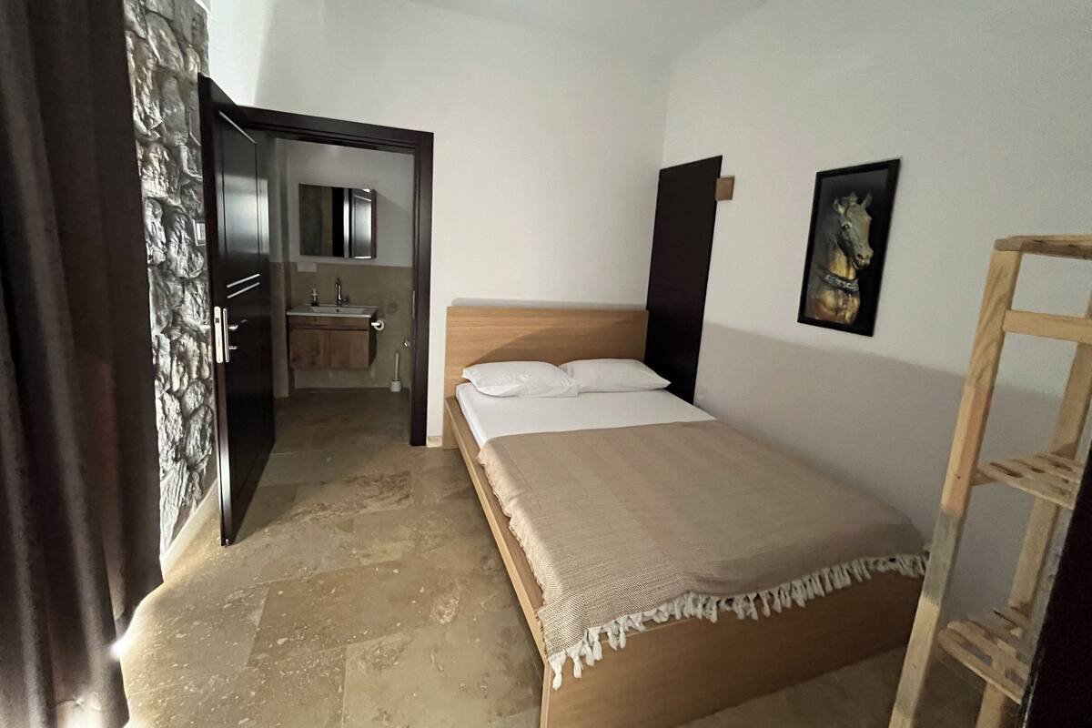 A bedroom with a bed, a wooden headboard, a wooden ladder, a bathroom with a mirror, and a stone wall.