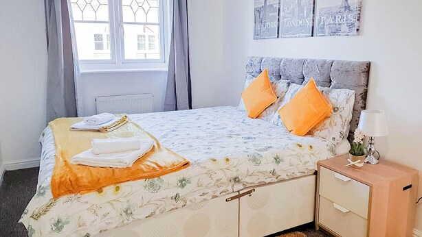 A neatly made bed with a floral bedspread and two orange pillows, a bedside table with a lamp, and a window with curtains. - Airport Lounge - 3Bed 6 Persons (Manchester)