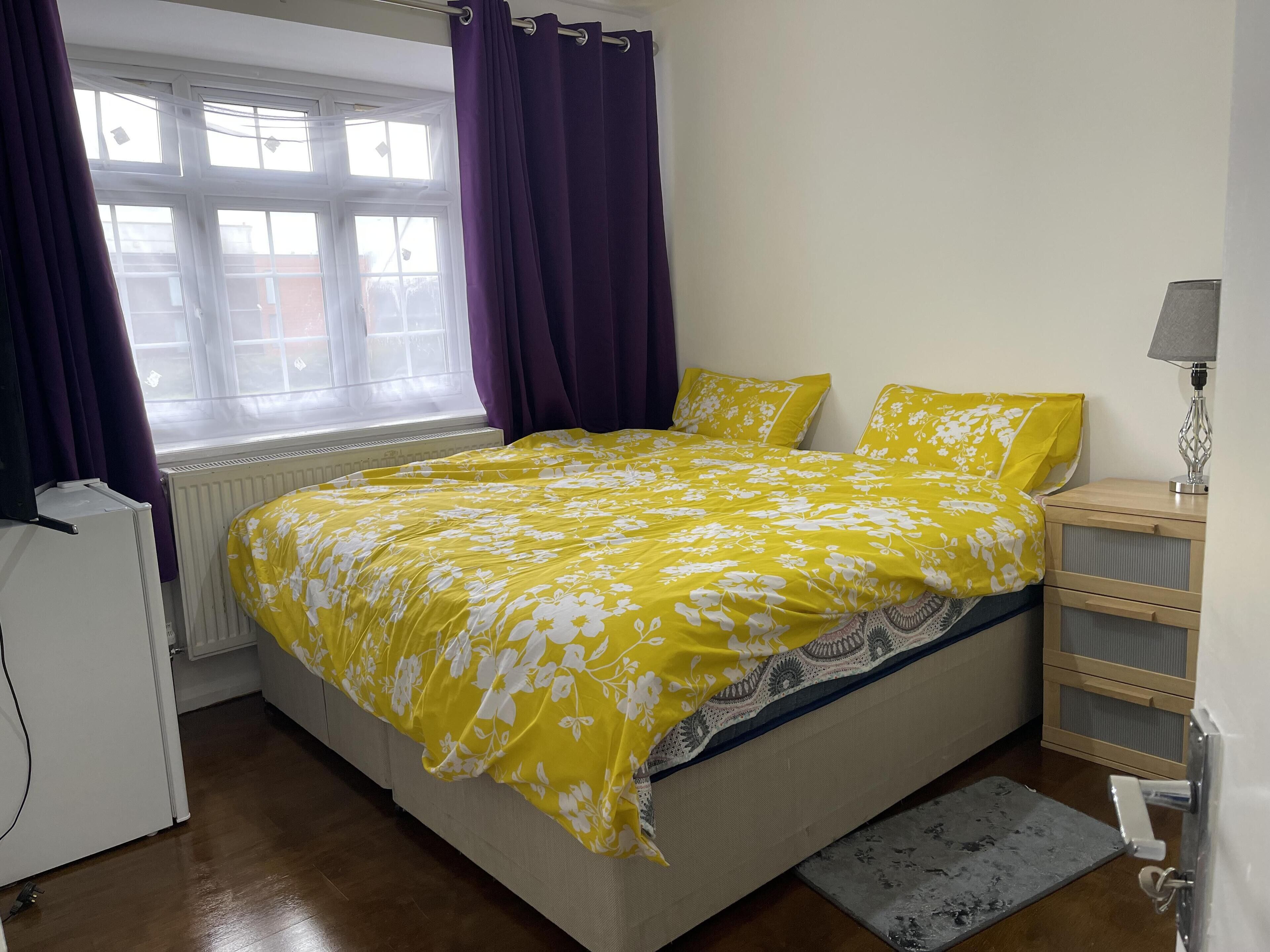 Quarto com cama, mesa de cabeceira, abajur, radiador e janela com cortinas.